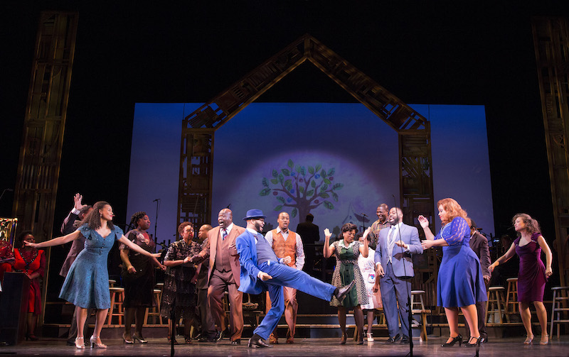 The 2016 cast of Cabin in the Sky. A man wearing a blue suit kicks his right leg out and his shoulders back